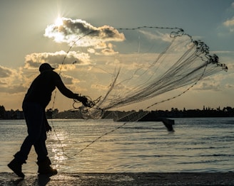 A silhouetted figure is casting a large fishing net over a calm body of water as the sun sets in the background. The sky is adorned with scattered clouds, and a hint of orange hues reflects on the water's surface. In the distance, the outline of trees and buildings can be seen along the horizon.