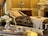 An elegant and luxurious interior featuring a vintage chaise lounge with ornate fabric and white lace throw. A decorative vase sits atop a polished wooden cabinet. In the foreground, a silver tray holds a delicate teacup and flowers, while a marble fireplace adds to the opulent atmosphere.