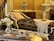 An elegant and luxurious interior featuring a vintage chaise lounge with ornate fabric and white lace throw. A decorative vase sits atop a polished wooden cabinet. In the foreground, a silver tray holds a delicate teacup and flowers, while a marble fireplace adds to the opulent atmosphere.