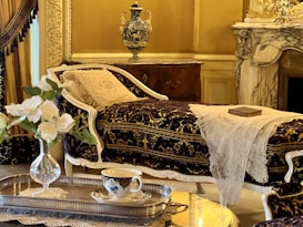An elegant and luxurious interior featuring a vintage chaise lounge with ornate fabric and white lace throw. A decorative vase sits atop a polished wooden cabinet. In the foreground, a silver tray holds a delicate teacup and flowers, while a marble fireplace adds to the opulent atmosphere.