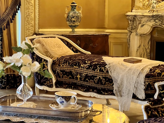 An elegant and luxurious interior featuring a vintage chaise lounge with ornate fabric and white lace throw. A decorative vase sits atop a polished wooden cabinet. In the foreground, a silver tray holds a delicate teacup and flowers, while a marble fireplace adds to the opulent atmosphere.