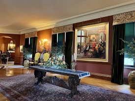 An elegant, well-lit room with ornate decorations includes a large painting depicting a historical or royal scene. The room features two high-backed chairs with yellow upholstery, a dark wooden table with a decorative table runner, and a floral arrangement. There are dark green drapes, a richly patterned rug, and several plants along the sides.