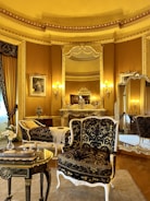 An elegant, luxurious room features antique furniture including ornate chairs with gold and black upholstery, an intricately carved table, and an elaborately decorated mantle. Large mirrors and rich gold curtains complement the opulent decor. Warm lighting from wall-mounted lamps enhances the grandeur.