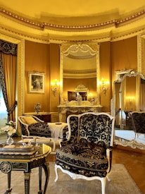An elegant, luxurious room features antique furniture including ornate chairs with gold and black upholstery, an intricately carved table, and an elaborately decorated mantle. Large mirrors and rich gold curtains complement the opulent decor. Warm lighting from wall-mounted lamps enhances the grandeur.