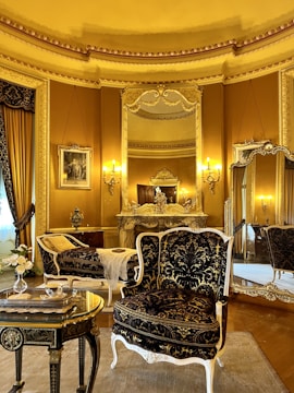 An elegant, luxurious room features antique furniture including ornate chairs with gold and black upholstery, an intricately carved table, and an elaborately decorated mantle. Large mirrors and rich gold curtains complement the opulent decor. Warm lighting from wall-mounted lamps enhances the grandeur.