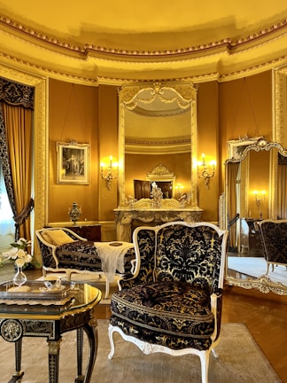 An elegant, luxurious room features antique furniture including ornate chairs with gold and black upholstery, an intricately carved table, and an elaborately decorated mantle. Large mirrors and rich gold curtains complement the opulent decor. Warm lighting from wall-mounted lamps enhances the grandeur.
