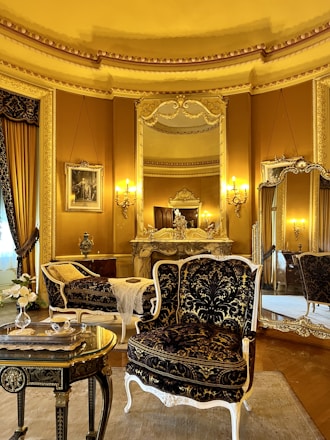 An elegant, luxurious room features antique furniture including ornate chairs with gold and black upholstery, an intricately carved table, and an elaborately decorated mantle. Large mirrors and rich gold curtains complement the opulent decor. Warm lighting from wall-mounted lamps enhances the grandeur.