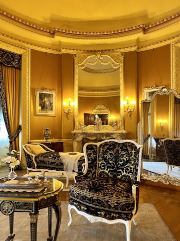 An elegant, luxurious room features antique furniture including ornate chairs with gold and black upholstery, an intricately carved table, and an elaborately decorated mantle. Large mirrors and rich gold curtains complement the opulent decor. Warm lighting from wall-mounted lamps enhances the grandeur.