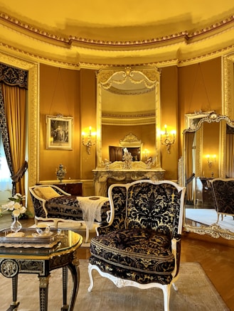 An elegant, luxurious room features antique furniture including ornate chairs with gold and black upholstery, an intricately carved table, and an elaborately decorated mantle. Large mirrors and rich gold curtains complement the opulent decor. Warm lighting from wall-mounted lamps enhances the grandeur.