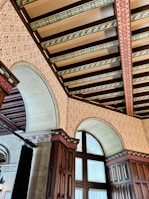 An ornate ceiling featuring intricate patterns and beams with decorative designs. The walls are adorned with detailed motifs, and arched windows with wooden frames are prominently displayed. The use of rich wood tones and elaborate details create a sense of elegance and historical grandeur.