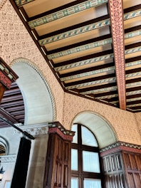 An ornate ceiling featuring intricate patterns and beams with decorative designs. The walls are adorned with detailed motifs, and arched windows with wooden frames are prominently displayed. The use of rich wood tones and elaborate details create a sense of elegance and historical grandeur.