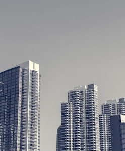 A skyline view with several modern high-rise buildings. The architecture features a mix of glass and concrete elements, with multiple balconies visible on the structures. The background is a clear sky with minimal clouds, giving the scene an airy and spacious feel.