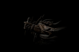 A dark, atmospheric scene featuring a figure cloaked in shadow with armor and a weapon. The intricate details of the armor include ropes and metal plates, casting a mysterious and intense aura.