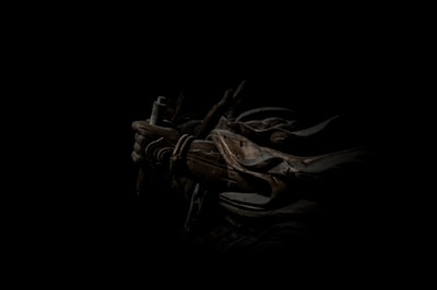 A dark, atmospheric scene featuring a figure cloaked in shadow with armor and a weapon. The intricate details of the armor include ropes and metal plates, casting a mysterious and intense aura.