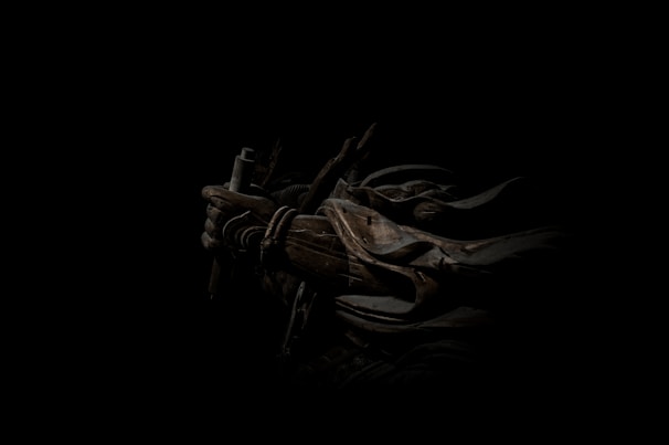 A dark, atmospheric scene featuring a figure cloaked in shadow with armor and a weapon. The intricate details of the armor include ropes and metal plates, casting a mysterious and intense aura.