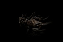 A dark, atmospheric scene featuring a figure cloaked in shadow with armor and a weapon. The intricate details of the armor include ropes and metal plates, casting a mysterious and intense aura.