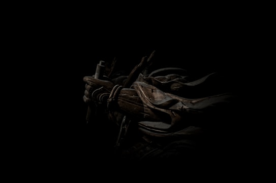 A dark, atmospheric scene featuring a figure cloaked in shadow with armor and a weapon. The intricate details of the armor include ropes and metal plates, casting a mysterious and intense aura.