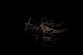 A dark, atmospheric scene featuring a figure cloaked in shadow with armor and a weapon. The intricate details of the armor include ropes and metal plates, casting a mysterious and intense aura.
