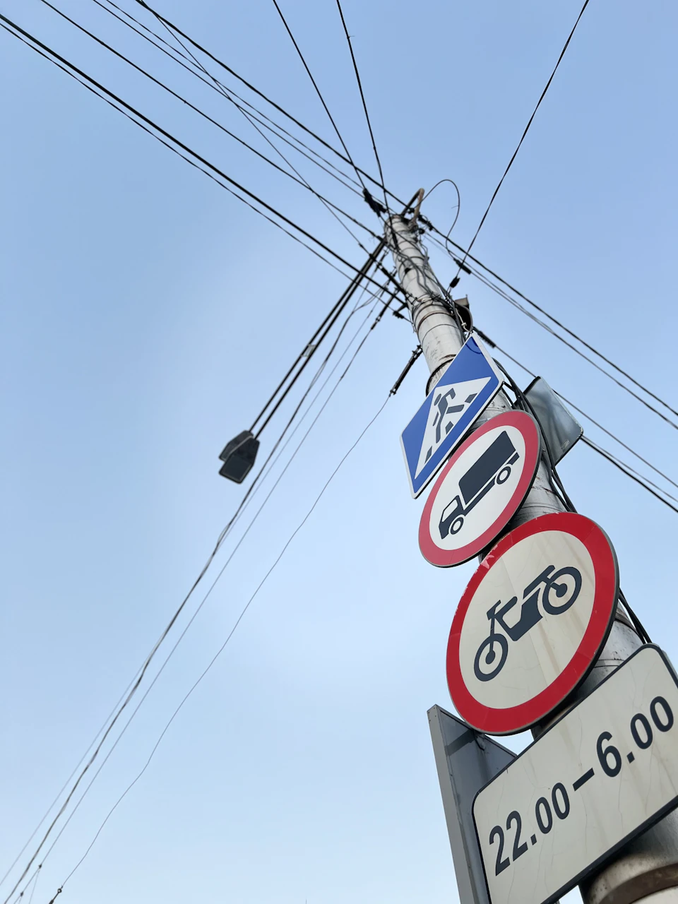 The image shows a street sign pole standing tall against a clear blue sky. The pole is adorned with three signs, each representing different types of traffic and directions. On the left side of the pole, there's a red and white "No Parking" sign indicating restricted parking zones. In the middle, two black and white road signs are visible - one reading "Motorcycles Allowed", suggesting that motorcycles can be driven in this area, while the other reads "Pedestrians Only", indicating that pedestrians should use designated crosswalks for their safety.

On the right side of the pole, there's a blue and white sign displaying the number "22:00" with an arrow pointing to the left, possibly indicating the time or direction of interest.