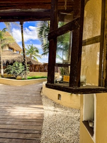 A tropical outdoor setting with a wooden pathway leading towards a thatched-roof building surrounded by lush palm trees. There is a window frame on the right side containing glass holders, and the ground is covered with white pebbles. The sky is clear with a few scattered clouds, adding to the tranquil atmosphere.