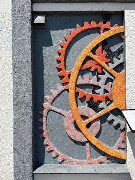 A textured wall features an artistic arrangement of oversized gears, primarily in shades of orange and gray. The gears are embedded in a recess, creating a sense of depth and industrial style. A small, rectangular plaque is visible to the right of the gears.