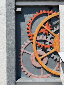 A textured wall features an artistic arrangement of oversized gears, primarily in shades of orange and gray. The gears are embedded in a recess, creating a sense of depth and industrial style. A small, rectangular plaque is visible to the right of the gears.