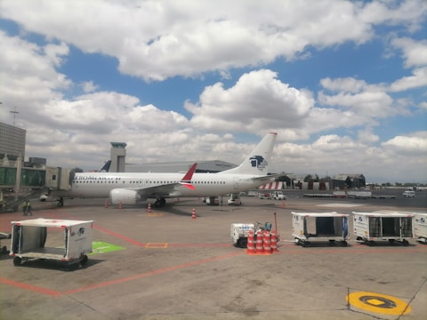 A white commercial airplane with Aeromexico branding is positioned on the tarmac at an airport. The plane is connected to a jet bridge for boarding. Several luggage carts and orange safety cones are visible nearby. The sky is partly cloudy, covering much of the blue background.