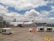 A white commercial airplane with Aeromexico branding is positioned on the tarmac at an airport. The plane is connected to a jet bridge for boarding. Several luggage carts and orange safety cones are visible nearby. The sky is partly cloudy, covering much of the blue background.