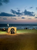 A serene beach scene at dusk with the sky transitioning from blue to pastel hues. A small, illuminated cabana with a light shines warmly on the sand, providing a cozy atmosphere. Few tables are set up near the cabana, while waves gently crash on the shore. A group of people can be seen in the background, enjoying the tranquil setting.