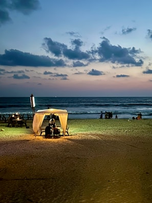 A serene beach scene at dusk with the sky transitioning from blue to pastel hues. A small, illuminated cabana with a light shines warmly on the sand, providing a cozy atmosphere. Few tables are set up near the cabana, while waves gently crash on the shore. A group of people can be seen in the background, enjoying the tranquil setting.