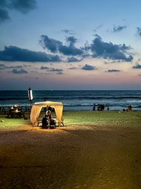 A serene beach scene at dusk with the sky transitioning from blue to pastel hues. A small, illuminated cabana with a light shines warmly on the sand, providing a cozy atmosphere. Few tables are set up near the cabana, while waves gently crash on the shore. A group of people can be seen in the background, enjoying the tranquil setting.