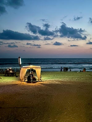 A serene beach scene at dusk with the sky transitioning from blue to pastel hues. A small, illuminated cabana with a light shines warmly on the sand, providing a cozy atmosphere. Few tables are set up near the cabana, while waves gently crash on the shore. A group of people can be seen in the background, enjoying the tranquil setting.