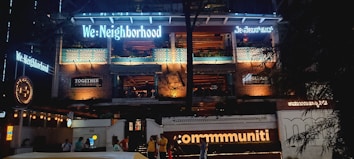 The image features the facade of a multi-story building illuminated with various lights. The building has signage with phrases like 'We: Neighborhood' and 'commmuniti' in large, bright letters, and there are signs of other businesses. The structure has exposed brickwork and balconies, with people visible at the entrance area.