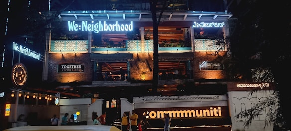 The image features the facade of a multi-story building illuminated with various lights. The building has signage with phrases like 'We: Neighborhood' and 'commmuniti' in large, bright letters, and there are signs of other businesses. The structure has exposed brickwork and balconies, with people visible at the entrance area.