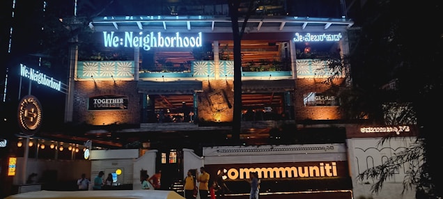 The image features the facade of a multi-story building illuminated with various lights. The building has signage with phrases like 'We: Neighborhood' and 'commmuniti' in large, bright letters, and there are signs of other businesses. The structure has exposed brickwork and balconies, with people visible at the entrance area.