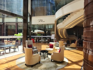 A modern, sophisticated lounge area with plush beige chairs accented with red cushions, arranged around small tables on a polished wooden floor. Large glass windows reveal an outside area with potted plants. The environment is well-lit with natural light streaming in. A curved staircase with sleek lines leads to an upper level. The ambiance is elegant and inviting, with soft lighting and stylish decor.