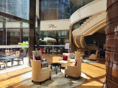 A modern, sophisticated lounge area with plush beige chairs accented with red cushions, arranged around small tables on a polished wooden floor. Large glass windows reveal an outside area with potted plants. The environment is well-lit with natural light streaming in. A curved staircase with sleek lines leads to an upper level. The ambiance is elegant and inviting, with soft lighting and stylish decor.