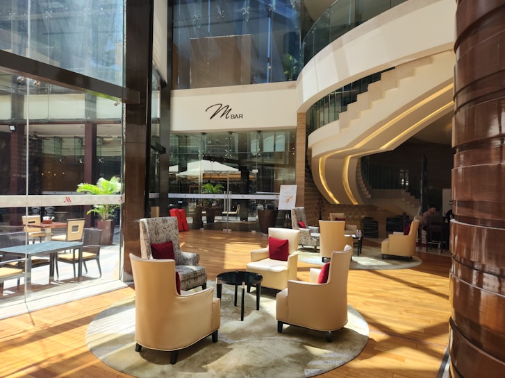 A modern, sophisticated lounge area with plush beige chairs accented with red cushions, arranged around small tables on a polished wooden floor. Large glass windows reveal an outside area with potted plants. The environment is well-lit with natural light streaming in. A curved staircase with sleek lines leads to an upper level. The ambiance is elegant and inviting, with soft lighting and stylish decor.