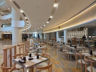 A modern and spacious dining area featuring wooden tables and chairs, elegant lighting, and a stylish interior design. The floor is made of polished stone tiles, and the ceiling has embedded lights in a sleek and curved layout. A bar area is visible in the background with stools and pendant lights.