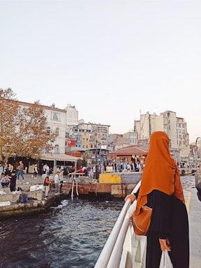 A woman wearing an orange hijab stands on a railing overlooking a vibrant waterfront scene. Numerous people are congregated along the water's edge and engaging in various activities. The background features a cluster of multi-story buildings with varying architectural styles and a few trees with autumn foliage. The sky is clear and bright.