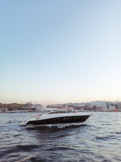 A sleek, modern yacht is gliding through a body of water with gentle waves. In the background, a cityscape featuring historic architecture, including minarets and domes, can be seen under a clear blue sky, creating a serene and picturesque scene.