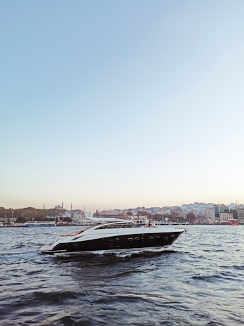 A sleek, modern yacht is gliding through a body of water with gentle waves. In the background, a cityscape featuring historic architecture, including minarets and domes, can be seen under a clear blue sky, creating a serene and picturesque scene.