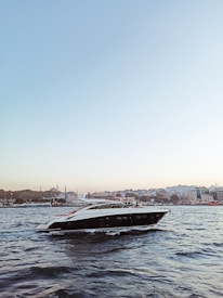 A sleek, modern yacht is gliding through a body of water with gentle waves. In the background, a cityscape featuring historic architecture, including minarets and domes, can be seen under a clear blue sky, creating a serene and picturesque scene.
