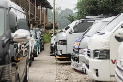 A row of parked vans and SUVs on either side of a narrow pathway, surrounded by greenery and rustic architecture. The vehicles are closely aligned and appear in various shades of white, silver, and blue.