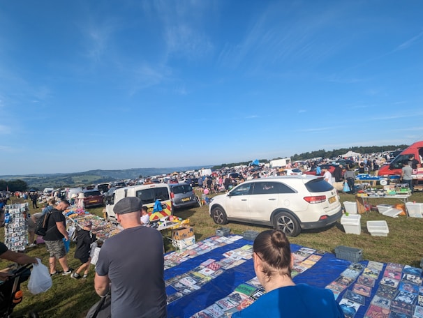 A busy outdoor market with numerous stalls displaying various goods, such as magazines and other items, set up on a grassy field. People are browsing the stalls, and several cars are parked nearby. The atmosphere appears lively and bustling, with clear blue skies overhead.