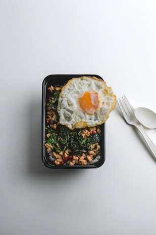 A black rectangular food container holds a meal consisting of cooked ground meat, dark green leafy vegetables, and slices of red chili peppers. A fried egg with a runny yolk is placed on top, adding contrast to the dish. Beside the container, there is a white plastic fork and spoon on a light gray surface.
