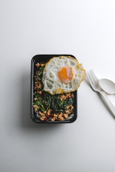 A black rectangular food container holds a meal consisting of cooked ground meat, dark green leafy vegetables, and slices of red chili peppers. A fried egg with a runny yolk is placed on top, adding contrast to the dish. Beside the container, there is a white plastic fork and spoon on a light gray surface.