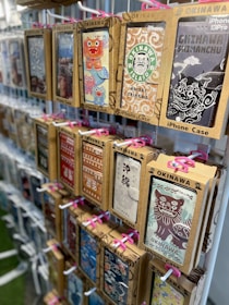 A display rack featuring iPhone cases decorated with various colorful and artistic designs. These designs include traditional Okinawan themes and motifs, such as Shisa (guardian lion dogs) and other local imagery. The cases are packaged in cardboard boxes, each with visible branding and labels related to Okinawa.