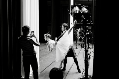 A dance performance taking place backstage, where a male dancer holds a female dancer in a lifted pose. A person in the foreground captures the moment with a smartphone. Stage lighting and equipment are visible in the scene.