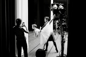 A dance performance taking place backstage, where a male dancer holds a female dancer in a lifted pose. A person in the foreground captures the moment with a smartphone. Stage lighting and equipment are visible in the scene.